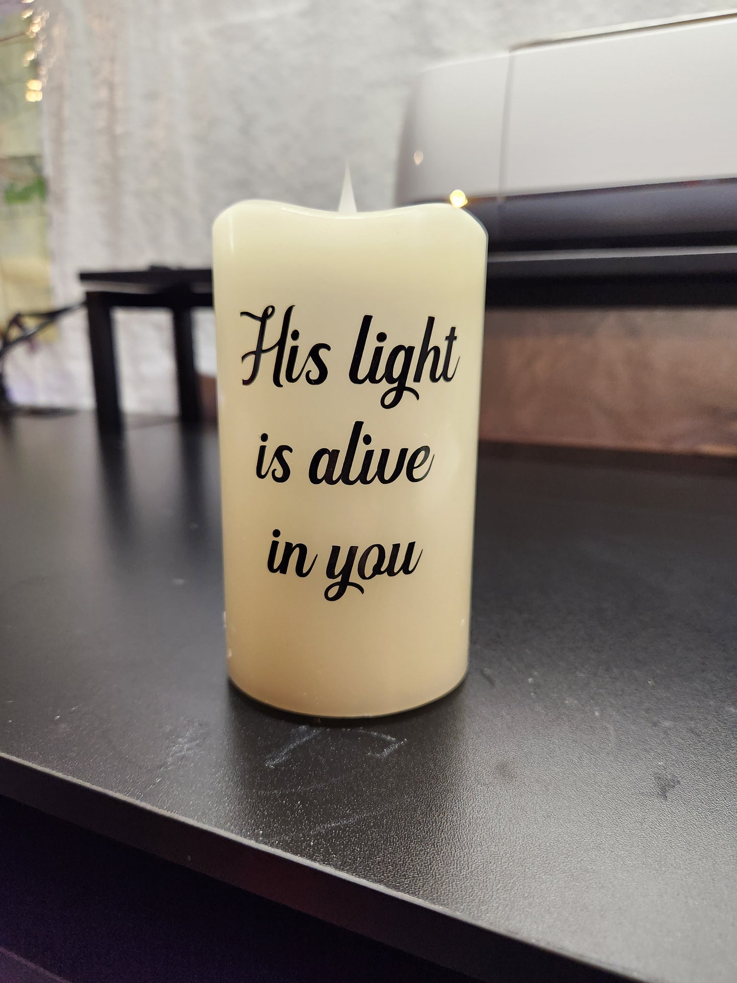 A beige memorial candle with black text that reads 'His light is alive in you'.