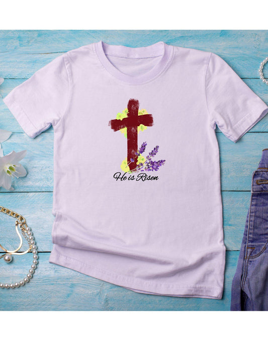 White t-shirt with a cross and 'He is Risen' text on a blue wooden surface